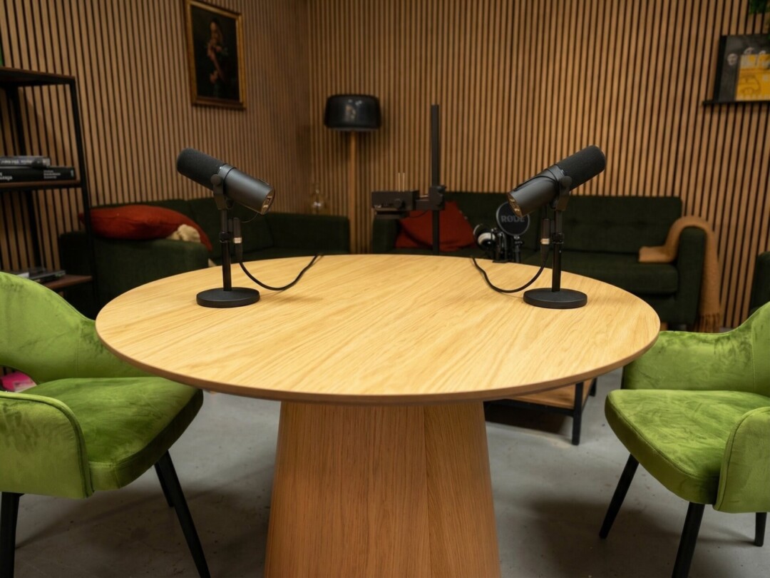 Professionelles Podcast-Setup mit zwei schwarzen Mikrofonen auf einem runden Holztisch im Podcast Studio Hannover, umgeben von grünen Samtsesseln und akustischen Holzpaneelen.