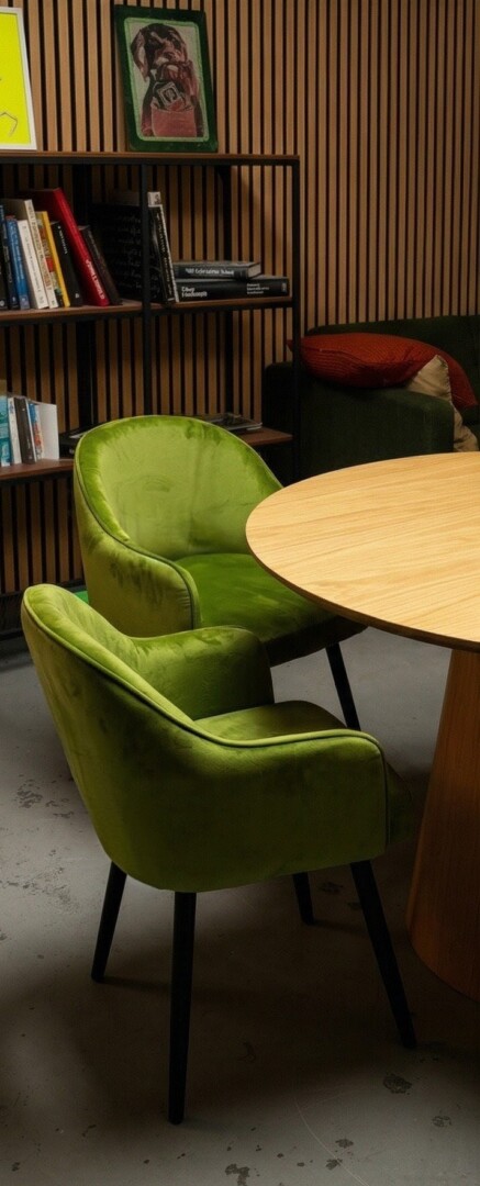 Zwei komfortable grüne Samtstühle an einem Aufnahmetisch neben einem Bücherregal, ideal für entspannte Interviews im Studio.